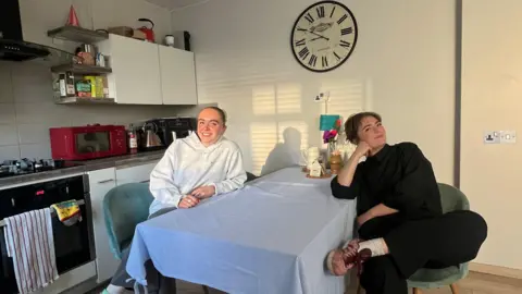 エラ・マーレイ エラ・マーレイとルームメイトのメイシーは、青いテーブルクロスがかかったキッチンのテーブルに座っています。二人とも微笑みます。このテーブルは、キッチンキャビネット、表面、コンロの組み合わせに近いものです。壁の一面には大きな掛け時計が掛けられています。